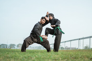 Asian male fighter wearing a pencak silat uniform with low stance pose and a hooded female fighter with one leg up and ready to kick in the outdoor background