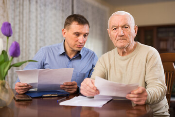 Obraz premium Senior man filling out papers with help of his adult son in living room at home