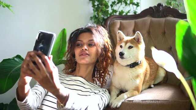 happy woman taking selfie photo with dog, sitting at home room spbd. Beautiful American female makes picture with pet and smiles while holding smartphone in hands in light interior. Attractive lady