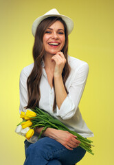 happy woman with flowers sitting on stool in front of yellow isolated back.