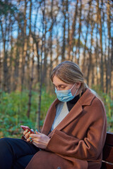 A charming girl in a medical protective mask with a phone in her hands is sitting on a bench in a Park