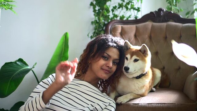 woman smiling and taking selfie photo with dog at home interior spbd. American African female hugs cute pet and takes picture, uses smartphone while sitting in modern interior with green plants. One