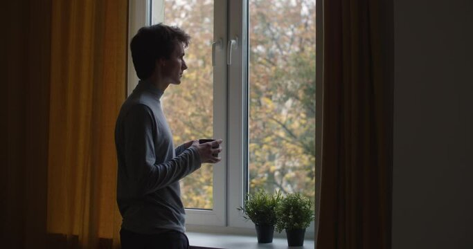 Pensive man standing by window indoors holding cup of tea slow motion. Back side portrait of sad guy drinking coffee alone at home background copy space text. Loneliness depression stress overcome