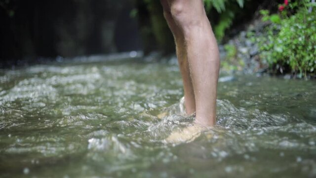 A male European hiking tourist is standing with bare feet in cold water creek a river close to a large waterfall cliff in the green forest jungle of Tanzania