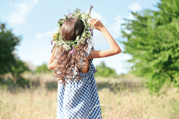 Little girl wearing wreath made of beautiful flowers in field on sunny day