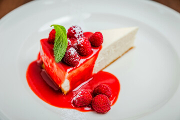 A piece of classic cheesecake new york, raspberry syrup topping, garnished with fresh berries and mint, white plate