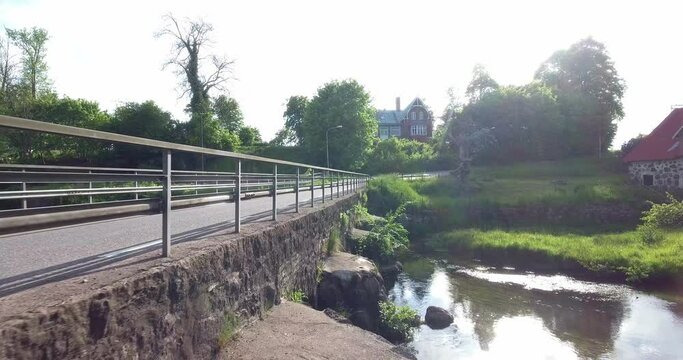 Car Driving On an Old Stone Bridge By a River Stream with Houses in Background, South Sweden Sk&aring;ne &Ouml;sterlen Svenstorp Aerial Low Forward Slow