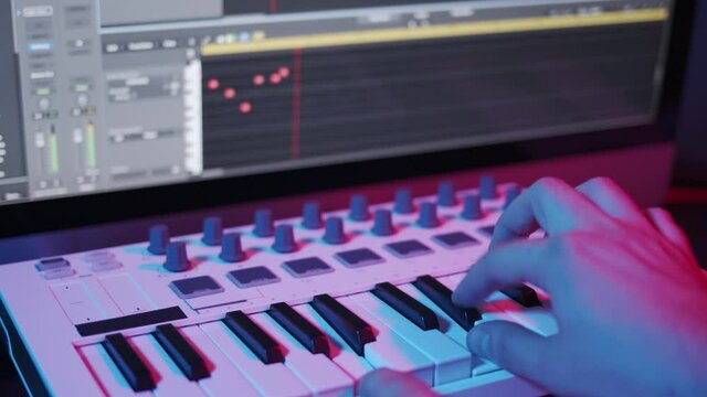Male hands recording music, playing electronic keyboard, midi keys on the table with neon lights. Closeup of male hands composing music in night, music sequencer and midi controller