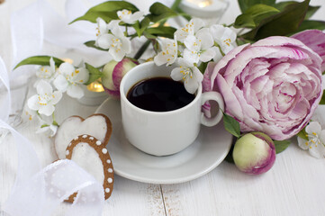 romantic breakfast: a bouquet of peonies and jasmine, a cup of coffee, heart-shaped cookies, a white ribbon on a light wooden background
