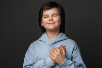 Cheerful boy 10-12 years old smiles gently and presses palms to heart. Positive emotions. Studio shot, gray background