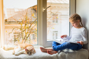 The child is looking at a book while sitting on the windowsill, and her pet is lying nearby. A...