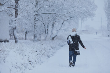 Naklejka na ściany i meble Man in dark clothes walks in the snow. A man walks along a snowy street in rural areas. A young, carelessly dressed guy travels in winter.