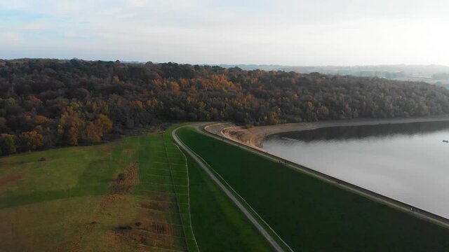 Forward drone aerial movement with autumn colours and dam of reservoir with walkway and walkers