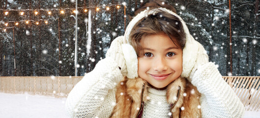 christmas, season and children concept - happy little girl wearing earmuffs over winter ice skating rink background - obrazy, fototapety, plakaty