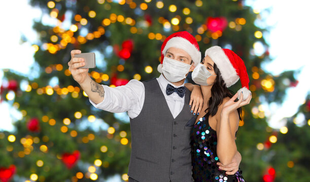 pandemic, health and winter holidays concept - couple wearing face protective medical masks for protection from virus disease taking selfie by smartphone over christmas tree lights on background