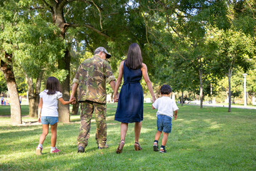 Obraz premium Rear view of Caucasian family holding hands and walking together in city park. Dad in camouflage uniform, long-haired mom and children enjoying holiday on nature. Family reunion and weekend concept