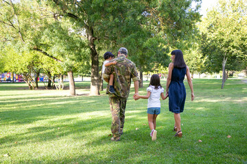 Back view of happy family walking together on meadow in park. Father wearing camouflage uniform,...