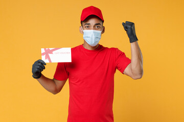 Delivery employee african man in red cap blank print t-shirt face mask gloves uniform work courier service on quarantine covid19 virus concept hold mockup coupon voucher isolated on yellow background