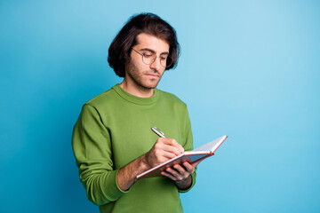 Photo of focused bearded guy hold notepad pen write wear spectacles green pullover isolated blue color background