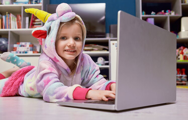 Little girl in pajama unicorn lyingat home on the wooden white floor with a laptop. Children's working with a laptop distance education. self-isolation