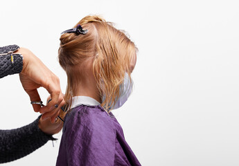 Fototapeta premium Hair cutting process. Beautiful little girl in medical mask on face at the barber shop, hairdresser making a hair style to cute caucasian 4-5 year old little girl