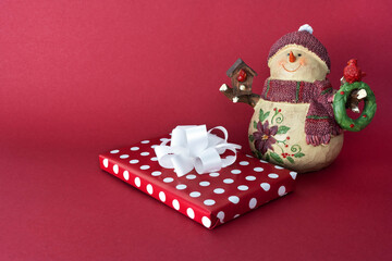 Christmas or New Year festive background. A red gift box with a white bow lies on a red background. On the right is a cute snowman in a red knitted hat and scarf