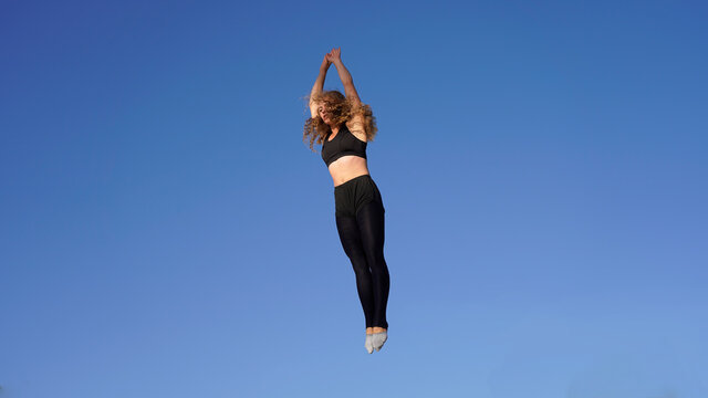 Beautiful girl jumping on a trampoline with hair fluttering in the wind against the background of the blue sky.