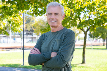 Happy mature man with crossed arms standing outside, looking at camera and smiling. Medium shot. Mature sporty person or active lifestyle concept