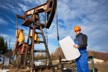 Worker holding plan of oil field and smiling to the camera at petroleum pump jack, wearing work...