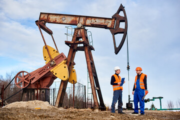 Two petroleum engineers in helmets and work vests standing on territory of oil field with oil well...