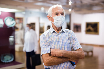 mature man in mask protecting against covid examines paintings on display in hall of art museum