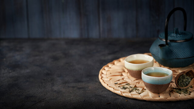 Asian food background banner with green tea, cups and teapot with leaves and free space for text on dark background.
