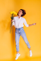 Studio shot on yellow background. Happy caucasian woman short hair wearing casual clothes, white shirt and denim pants, holding bouquet of yellow asters.  