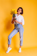 Studio shot on yellow background. Happy caucasian woman short hair wearing casual clothes, white shirt and denim pants, holding bouquet of yellow asters.  