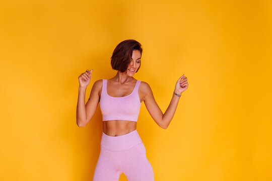 Sports woman stands on yellow background, demonstrating her abs, satisfied with the results of fitness training and diet, has a happy facial expression, wears a sports top and tight leggings  