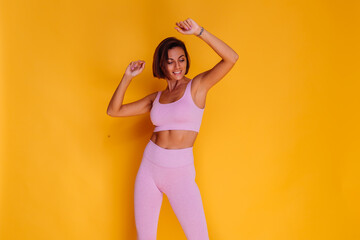 Sports woman stands on yellow background, demonstrating her abs, satisfied with the results of fitness training and diet, has a happy facial expression, wears a sports top and tight leggings  