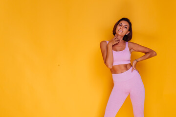 Sports woman stands on yellow background, demonstrating her abs, satisfied with the results of fitness training and diet, has a happy facial expression, wears a sports top and tight leggings  
