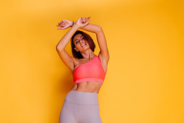 Sport and yoga lifestyle concept. Young fit pretty strong woman dressed in sport clothes, stylish top and leggings, poses against yellow background. Studio shot.   