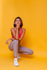 Sport and yoga lifestyle concept. Young fit pretty strong woman dressed in sport clothes, stylish top and leggings, poses against yellow background. Studio shot.   
