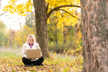 Cute woman with laptop in the autumn park. Beauty nature scene with colorful foliage background, yellow trees and leaves at fall season. Autumn outdoor lifestyle. Happy smiling woman on fall leaves