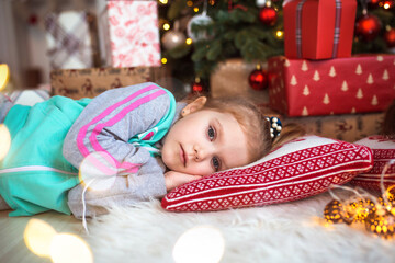 A little girl in home clothes is lying under the Christmas tree near the boxes with gifts on the pillow and dreams. Lights of garlands, the night before Christmas, new year.