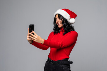 Portrait of a pretty woman dressed in red christmas hat holding mobile phone while standing and looking at camera isolated over gray background