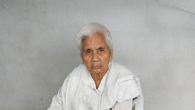 Asian old woman looking to camera with serious face isolated concrete grey wall background. Rural lifestyle concept.