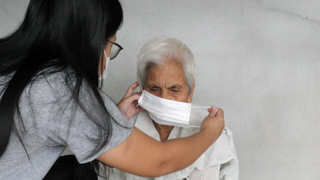 Asian Grandchild Woman Wearing A White Face Mask To Old Grandmother Prevent From Covid 19. Isolated Concrete Grey Wall Background. Rural Lifestyle And Health Care Concept.