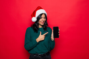 Smiling young woman in santa claus hat pointing finger at blank screen smartphone over red background