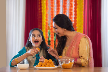 Daughter eating ladoo while her mother watches her	