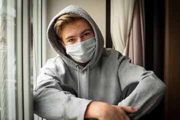Sad teenage boy confined at home by the coronavirus crisis, looks bored out the window without being able to leave home, defocused background.