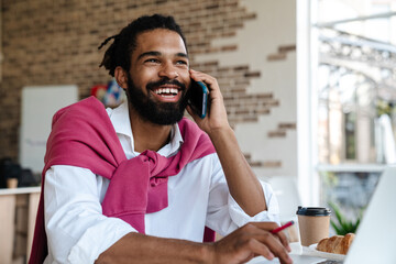 Smiling african american guy talking on cellphone and working with laptop