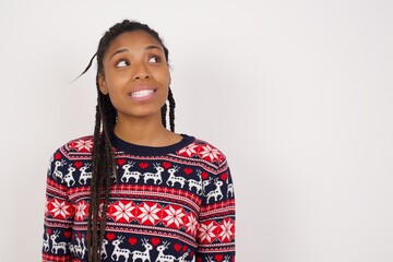 Oops! Portrait of Young beautiful African American woman wearing Christmas sweater against white wall,   clenches teeth and looks confusedly aside, realizes her bad mistake,