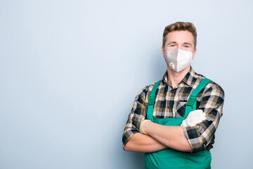 Portrait of expert handyman wearing green overalls white respiratory face mask standing with folded hands isolated on gray background with blank space